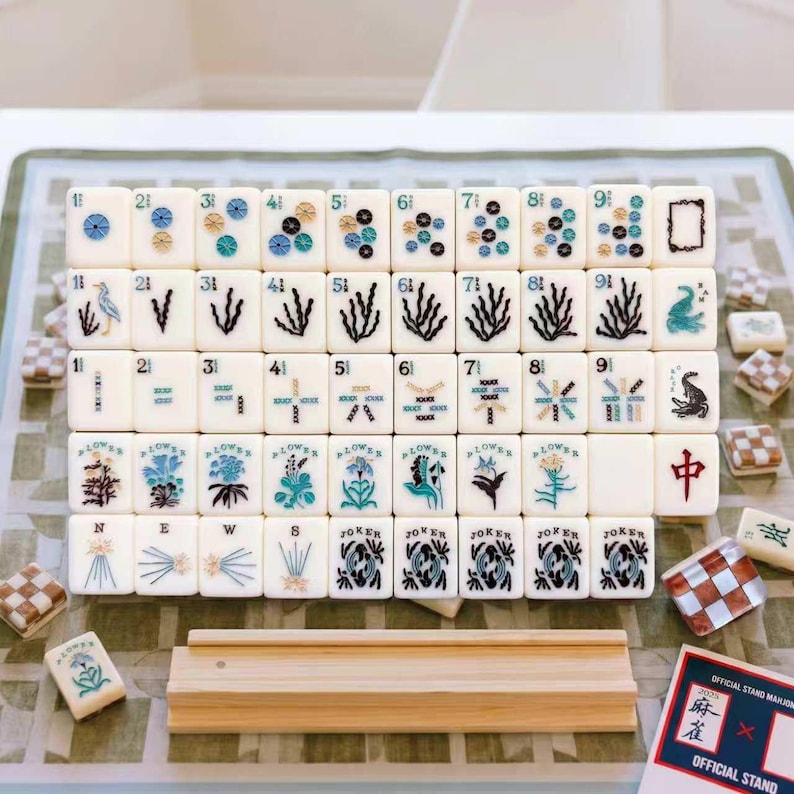 May include: A set of white Mahjong tiles featuring blue and black botanical, numerical, and character designs. The tiles are arranged in a grid with a wooden rack in front and a small card labeled 2025 Official Stand. Some tiles show floral patterns, birds, and the word NEWS.