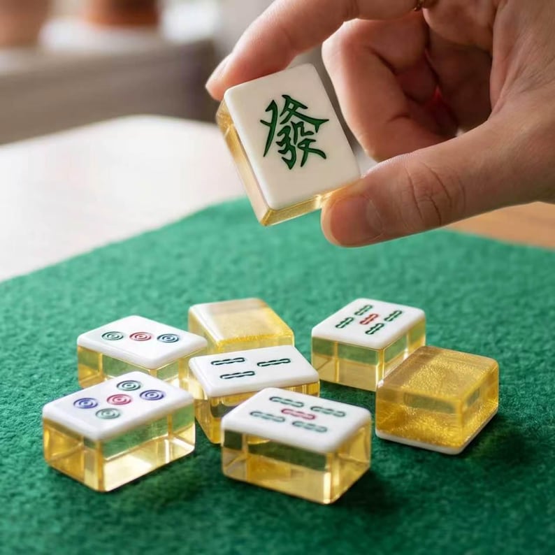 May include: A set of rectangular Mahjong tiles featuring white tops with green and red engraved symbols and translucent gold-tinted bases. One tile is held up, displaying a green Chinese character, while others rest on a green felt surface.