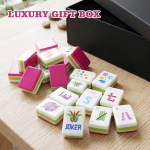 May include: A set of mahjong tiles featuring white faces with colorful floral, numerical, and joker designs, paired with magenta and green sides. The tiles are displayed alongside a large black box labeled LUXURY GIFT BOX in white text.