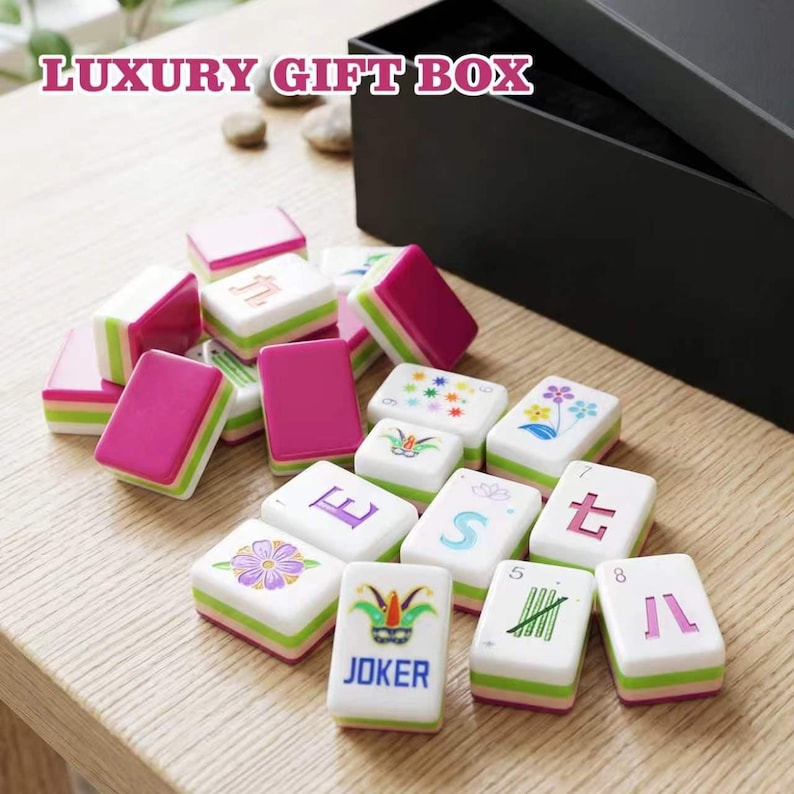 May include: A set of mahjong tiles featuring white faces with colorful floral, numerical, and joker designs, paired with magenta and green sides. The tiles are displayed alongside a large black box labeled LUXURY GIFT BOX in white text.