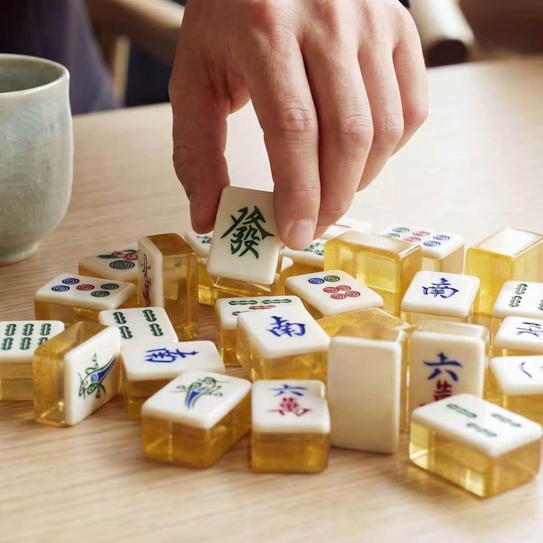 May include: A collection of rectangular Mahjong tiles with white tops featuring various green, blue, and red engraved symbols and characters, and translucent amber-colored bases. One tile is held upright, displaying a green Chinese character.