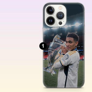 May include: A clear smartphone case featuring a printed image of a soccer player in a white jersey holding a large silver trophy on a field. The design includes the player looking over their shoulder with the stadium lights visible in the background.