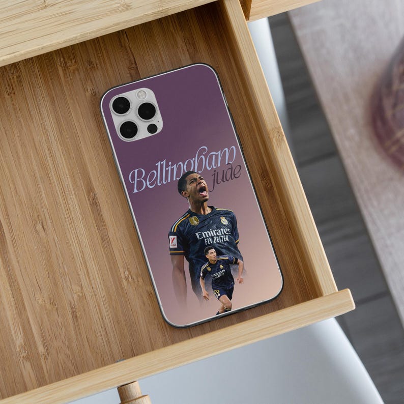 May include: A smartphone case with a purple and light pink gradient background featuring a graphic of a soccer player in a dark blue jersey. The text Bellingham Jude is printed in a white script font above the player, who is shown in two poses.