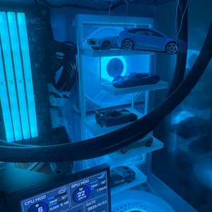 Peut inclure: Un intérieur de boîtier d'ordinateur éclairé par des LED bleues, comprenant une étagère blanche verticale présentant cinq voitures miniatures. Un écran numérique affiche les statistiques du système, dont le CPU, le GPU, la RAM et le HDD.