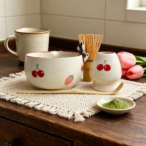 May include: A ceramic matcha set featuring a white bowl with red cherry and strawberry illustrations, a bamboo whisk with a small dog figurine, a bamboo scoop, a white whisk holder with a cherry print, and a small dish of green matcha powder.