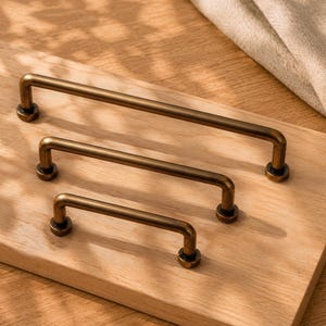 Può includere: Tre maniglie per armadietti in metallo con finitura bronzo scuro, disposte in ordine di grandezza decrescente. Ogni maniglia presenta un design semplice a barra dritta con due supporti cilindrici alle estremità, per un'estetica pulita e funzionale.
