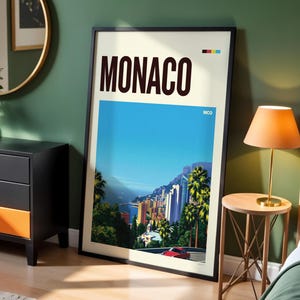 May include: A framed vertical poster featuring the text MONACO in large dark brown letters at the top, with a vibrant illustration of the Monaco coastline, palm trees, and a red car below, plus the text MCO and a small color block graphic.