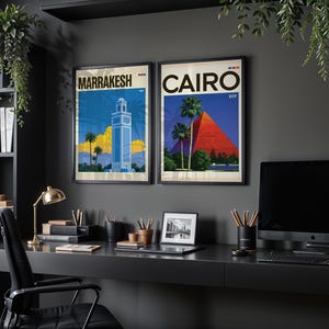 May include: Two framed travel posters hanging on a dark wall. The left poster features a blue tower in Marrakesh with palm trees and yellow clouds. The right poster depicts the red pyramids of Cairo with palm trees and the text CAIRO EGY.