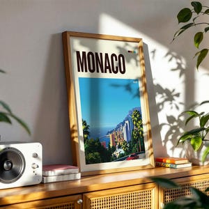 May include: A framed vertical travel poster featuring the word MONACO in bold dark brown letters at the top. The artwork depicts a vibrant coastal cityscape with palm trees, colorful buildings, a blue sea, and a red car on a winding road.