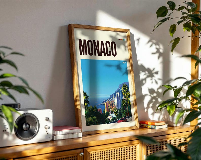 May include: A framed vertical travel poster featuring the word MONACO in bold dark brown letters at the top. The artwork depicts a vibrant coastal cityscape with palm trees, colorful buildings, a blue sea, and a red car on a winding road.