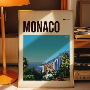 May include: A framed travel poster featuring the word MONACO in large black letters at the top. The artwork depicts a coastal cityscape with palm trees, high-rise buildings, a blue sea, and a red car on a road. The text MCO appears in the corner.