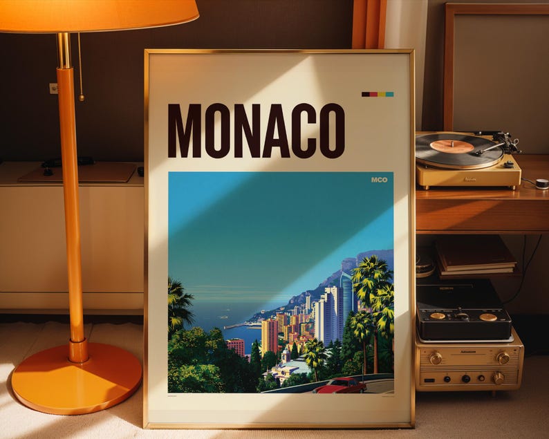 May include: A framed travel poster featuring the word MONACO in large black letters at the top. The artwork depicts a coastal cityscape with palm trees, high-rise buildings, a blue sea, and a red car on a road. The text MCO appears in the corner.