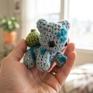 May include: A small, light blue crocheted creature with a textured surface, featuring dark, glossy eyes, teal-colored diamond-shaped patches on its body, and a rounded, olive-green bulb-like structure attached to its back, crafted in a soft yarn.