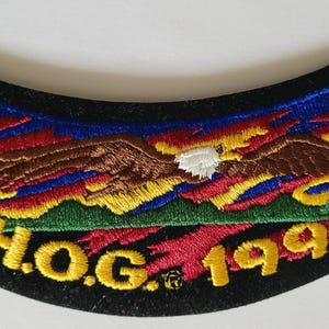 May include: A curved embroidered patch featuring a brown eagle with a white head flying over a green landscape against a blue sky with red and yellow flames. The text H.O.G. 1996 is stitched in yellow thread along the bottom edge of the patch.