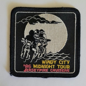 May include: A square embroidered patch with a black background featuring a large beige moon, clouds, and two motorcyclists. The text reads WINDY CITY '86 MIDNIGHT TOUR JERSEYPINE CRUISERS in yellow and pink thread with a white border and black edge.