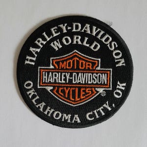 May include: A circular black embroidered patch featuring the white text HARLEY-DAVIDSON WORLD and OKLAHOMA CITY, OK. In the center is an orange and white Harley-Davidson Motor Cycles logo with the text MOTOR HARLEY-DAVIDSON CYCLES.
