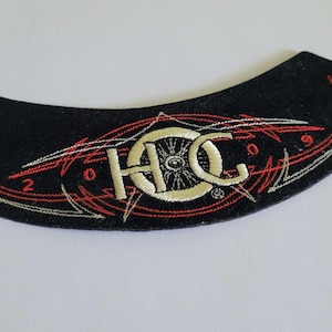 May include: A curved black fabric patch featuring embroidered details in cream and red thread. The design includes the letters HOG, a central wheel graphic, and the numbers 20 and 09 flanking the central emblem with thin red and cream lines.