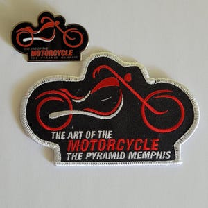 May include: A black and red embroidered patch and a matching metal pin, both featuring a stylized motorcycle silhouette and the text THE ART OF THE MOTORCYCLE THE PYRAMID MEMPHIS. The patch has a white stitched border and the pin has a metallic finish.