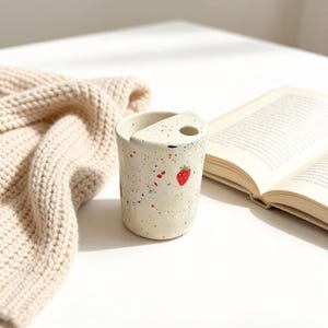 May include: A cylindrical ceramic cup with a cream-colored base, featuring a speckled pattern of red, blue, and yellow dots. The cup has a unique semi-circular top rim with a small circular opening and a painted red strawberry design on the side.