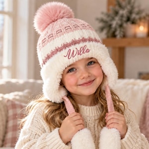 May include: A knit winter hat featuring a pink and white color scheme with a large pink pom-pom on top. The hat has ear flaps with long braided ties ending in white pom-poms. The name Willa is embroidered in pink script on the front.