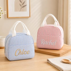 Puede incluir: Dos bolsas térmicas para el almuerzo con asas blancas y un patrón de rayas verticales. Una bolsa es azul claro con el nombre Chloe bordado en dorado, y la otra es rosa claro con el nombre Rachel bordado en letras cursivas blancas.