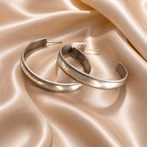 May include: A pair of silver-toned hoop earrings featuring a smooth, polished center band flanked by two thin, textured beaded edges. The earrings have a curved, semi-circular design with a post-back closure, showcasing a metallic finish.