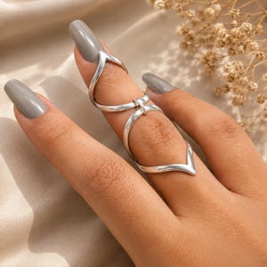 May include: A silver-toned full-finger ring featuring a segmented design with two V-shaped ends connected by a central hinge. The metal band wraps around the finger, with one V-point positioned near the nail and the other near the knuckle.