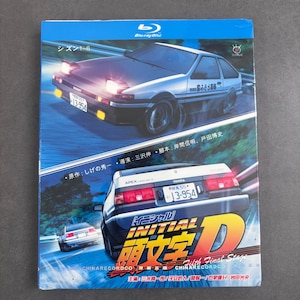 Pode incluir: Uma caixa de Blu-ray de Initial D com um carro esportivo Toyota AE86 branco e preto em movimento em uma estrada de montanha. A capa exibe o carro de ângulos frontais e traseiros, texto em japonês e o título Initial D em letras amarelas grandes.