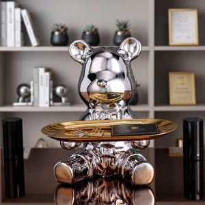 Peut inclure: Une figurine en forme d'ours argenté et réfléchissant tenant un plateau ovale doré horizontal. Le plateau contient un stylo doré, deux pinces à dessin en métal argenté et une petite carte rectangulaire noire avec du texte doré.