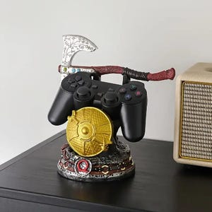 Peut inclure: Une manette de jeu vid&eacute;o noire repose sur un support d&eacute;coratif comprenant un bouclier circulaire dor&eacute;, une hache argent&eacute;e aux gravures complexes et une base orn&eacute;e d'un symbole Q rouge, con&ccedil;u pour maintenir la manette en toute s&eacute;curit&eacute;.