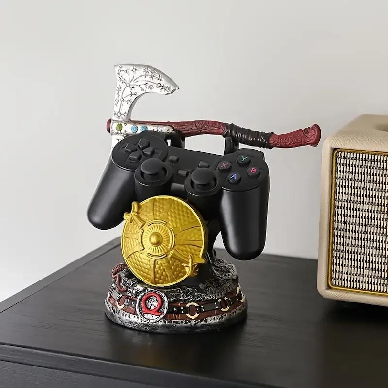 Peut inclure: Une manette de jeu vid&eacute;o noire repose sur un support d&eacute;coratif comprenant un bouclier circulaire dor&eacute;, une hache argent&eacute;e aux gravures complexes et une base orn&eacute;e d'un symbole Q rouge, con&ccedil;u pour maintenir la manette en toute s&eacute;curit&eacute;.