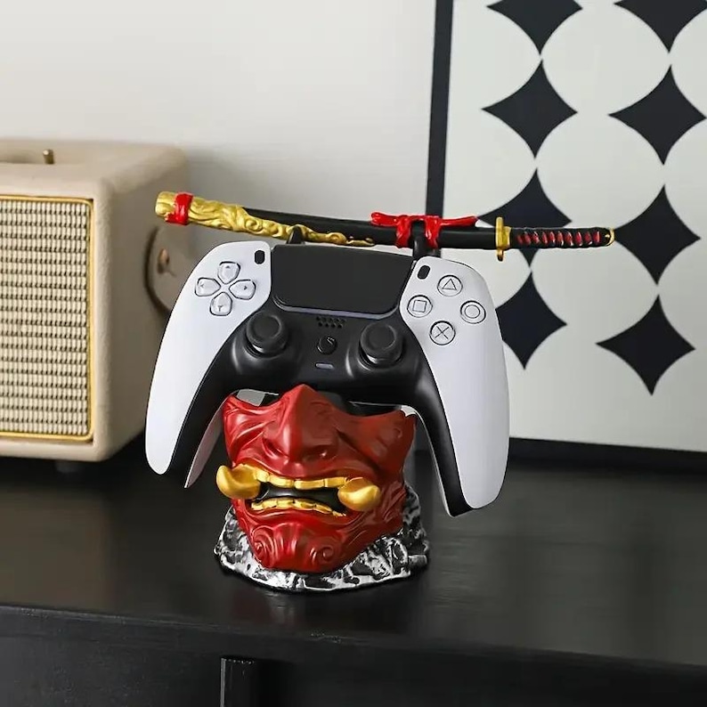 Peut inclure: Un support de manette de jeu avec une base en forme de masque de samoura&iuml; rouge et dor&eacute;, surmont&eacute; d'un katana miniature pos&eacute; horizontalement. Le support maintient fermement une manette de jeu vid&eacute;o sans fil blanche et noire.