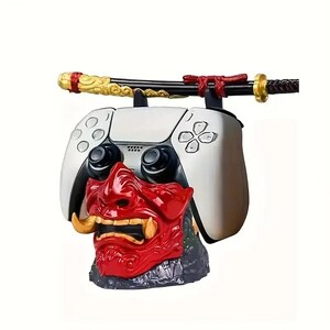 Peut inclure: Un support de manette de jeu vid&eacute;o blanc avec une base en forme de masque oni rouge et dor&eacute; aux crocs saillants. Un katana noir et dor&eacute; repose horizontalement sur le dessus de la manette, maintenue par deux supports noirs.