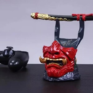 Peut inclure: Un support de manette en forme de masque oni rouge et noir avec des crocs et des cornes dor&eacute;s, con&ccedil;u pour maintenir une manette de jeu noire. Le support comprend un accessoire horizontal noir et dor&eacute; rappelant une &eacute;p&eacute;e pos&eacute; sur le dessus.