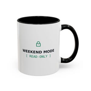 May include: A white ceramic mug with a black handle and interior. The front features a green padlock icon above the black text WEEKEND MODE and the green text [ READ-ONLY ]. The mug has a cylindrical body and a curved handle on the right side.