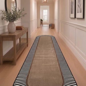 May include: A long, oval-shaped runner rug featuring a solid tan center bordered by a thick brown band and an outer edge decorated with a black and white striped pattern. The rug is positioned on a light wood floor in a hallway with white walls.