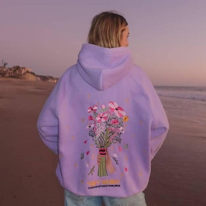 Op de afbeelding: Een lichtpaarse hoodie met op de achterkant een geborduurd boeket van roze, witte en gele bloemen. Het ontwerp bevat kleine gele steraccenten en de tekst "GIFT GIVING" in het geel gedrukt onder het bloemstuk.