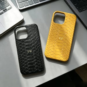 Puede incluir: Dos fundas rectangulares para smartphone con un patrón texturizado de piel de serpiente. Una funda es negra con las iniciales doradas FW grabadas en el centro, y la otra es amarilla con las iniciales doradas LJ grabadas en el centro.