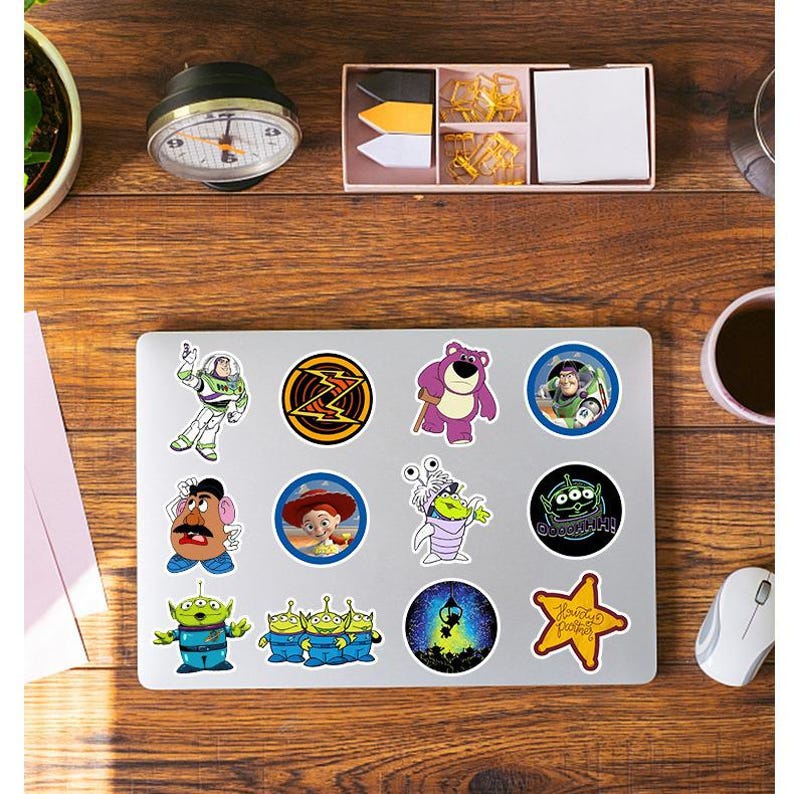 May include: A silver laptop decorated with twelve Toy Story character stickers, including Buzz Lightyear, Lotso, Mr. Potato Head, Jessie, and the green aliens. The stickers feature various circular, star, and die-cut shapes with colorful designs.