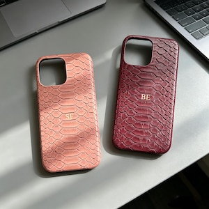 Puede incluir: Dos fundas de teléfono rectangulares con un patrón de piel de serpiente texturizado. La funda de la izquierda es de color rosa claro con las letras doradas SF en relieve en el centro. La derecha es de color burdeos con las letras BE.