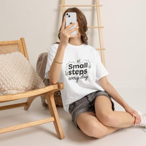 May include: A white short-sleeve t-shirt featuring the black text Small steps every day surrounded by a dotted path and small green leaf illustrations. The shirt has a crew neckline and a standard fit, worn with dark gray denim shorts.