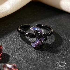 Puede incluir: Un anillo de metal negro con un diseño de media luna con acabado iridiscente en morado y azul. La banda tiene una superficie texturizada y ondulada con un diseño abierto. El texto Solac Jewelry Co es visible en la esquina inferior derecha.