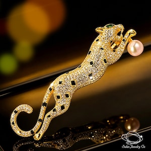 May include: A gold-toned leopard brooch encrusted with clear crystals and black enamel accents. The leopard is depicted in a leaping pose, featuring a single green crystal eye and holding a round, pale pink pearl between its front paws.