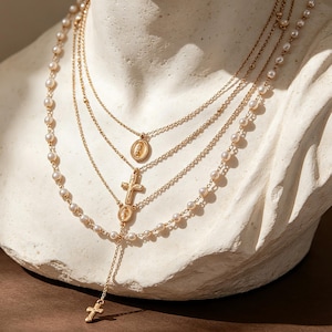 May include: A layered gold-tone necklace set featuring a strand of small white pearls and three delicate gold chains. The chains are adorned with two small oval religious pendants and two gold crosses, creating a tiered, multi-strand design.