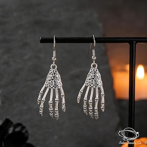 Peut inclure: Une paire de boucles d'oreilles pendantes argentées en forme de mains squelettiques. Chaque boucle présente une paume osseuse détaillée et des doigts articulés suspendus à un crochet simple. Le métal a une finition oxydée sombre.