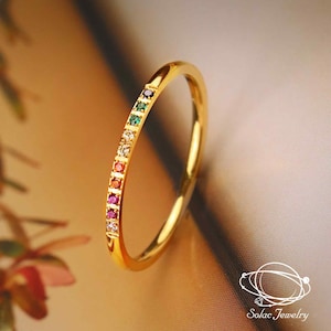 Op de afbeelding: Een dunne gouden ring met een rij kleine, veelkleurige edelstenen langs de band. De stenen hebben tinten van donkerpaars, groen, lichtblauw, transparant, oranje, rood en roze. De tekst Solac Jewelry is zichtbaar in de onderste hoek van de afbeelding.