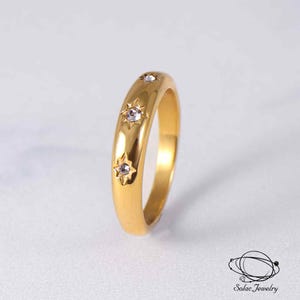 May include: A gold-toned band ring featuring three embedded clear stones, each set within a star-shaped engraving. The ring has a smooth, polished finish and a rounded profile. The text Solac Jewelry is visible in the bottom right corner.