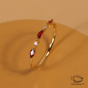 May include: A gold ring featuring a delicate band set with alternating marquise-cut red stones and small round clear stones. The jewellery piece includes a small logo and the text Solac Jewelry Co in the bottom right corner of the frame.