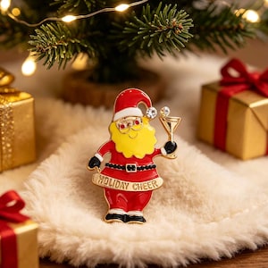 May include: A gold-tone metal brooch shaped like Santa Claus wearing a red suit and hat, a yellow beard, and glasses. Santa holds a martini glass with a clear crystal accent and a banner at the bottom with the text HOLIDAY CHEER in gold letters.