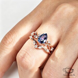 May include: A rose gold ring set featuring a pear-shaped, color-changing alexandrite center stone flanked by small clear accent stones. The bands are designed with a delicate leaf-like motif. The text Solac Jewelry Co is visible in the corner.